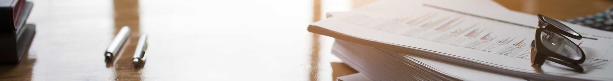 stack of reports on a corporate desk with pens and eyeglasses nearby